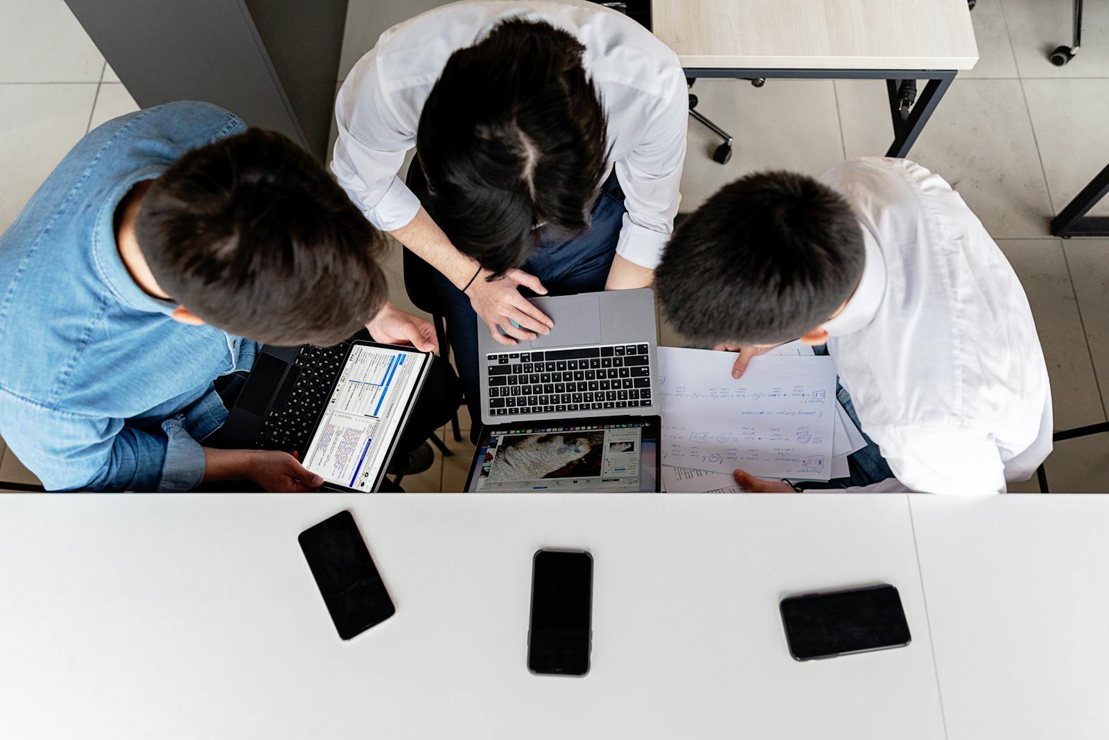 Three colleagues working together on laptops at an office desk with documents.