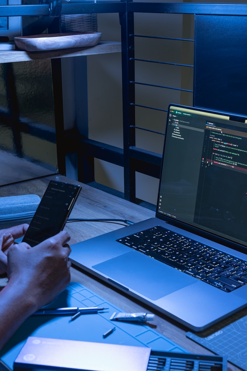 Person using smartphone next to laptop with code on screen.