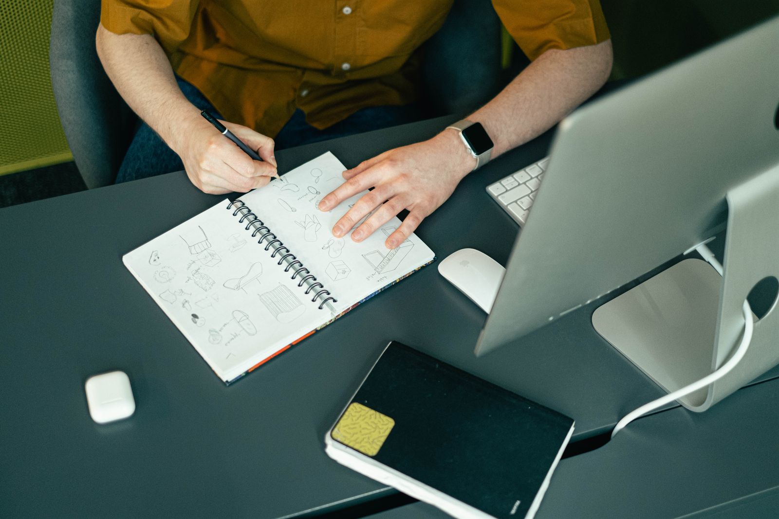 A person sketching in a notebook at a modern office workspace with a computer and accessories.