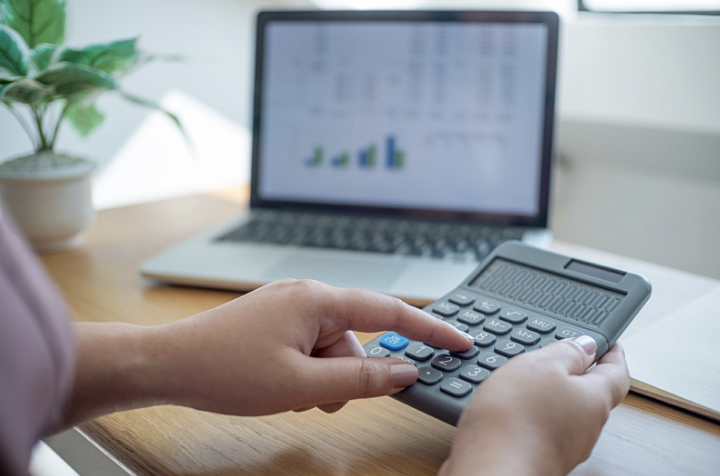 A person using a calculator beside a laptop displaying business data, representing the budgeting and planning involved in building an effective web application. The image reflects cost estimation, resource management, financial planning, and informed decision-making during web application development.